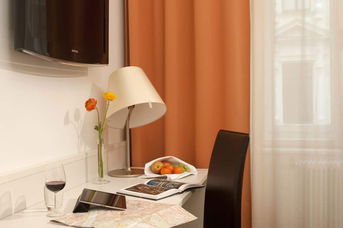 A hotel room with a flat-screen TV, a table with a lamp, a vase with flowers, a bowl of fruit, a wine glass, and a map.