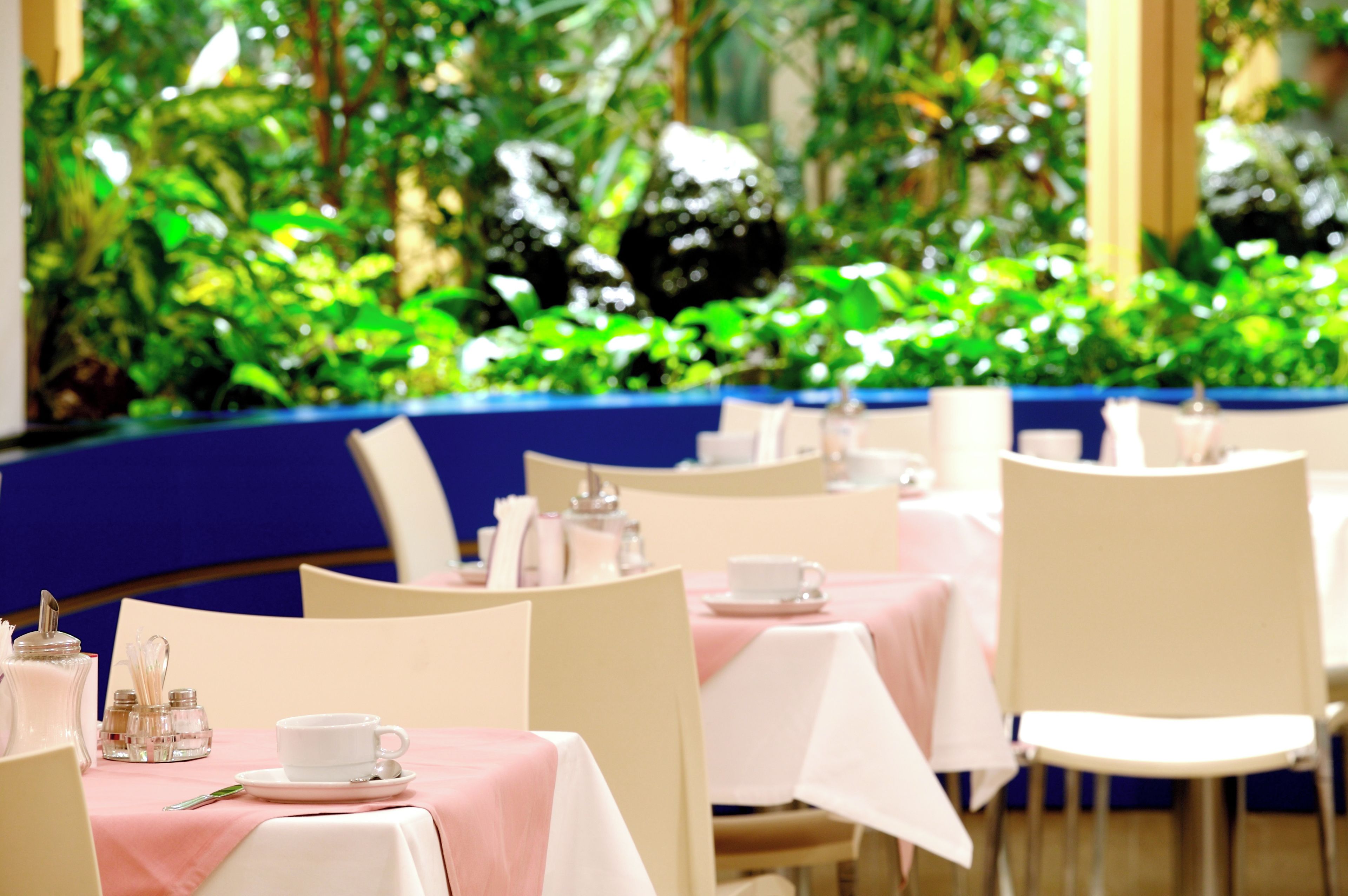 Ein Restaurant mit mit weißen Tischdecken gedeckten Tischen und Stühlen, vor einer Kulisse aus üppigem Grün.