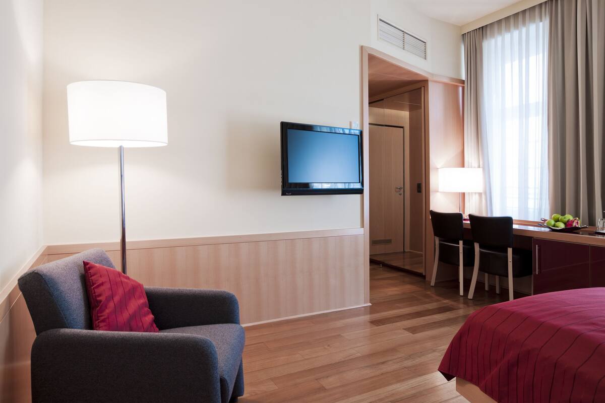 A hotel room with a flat-screen TV mounted on the wall, a desk with two chairs, a bed with a red bedspread, and a floor lamp.