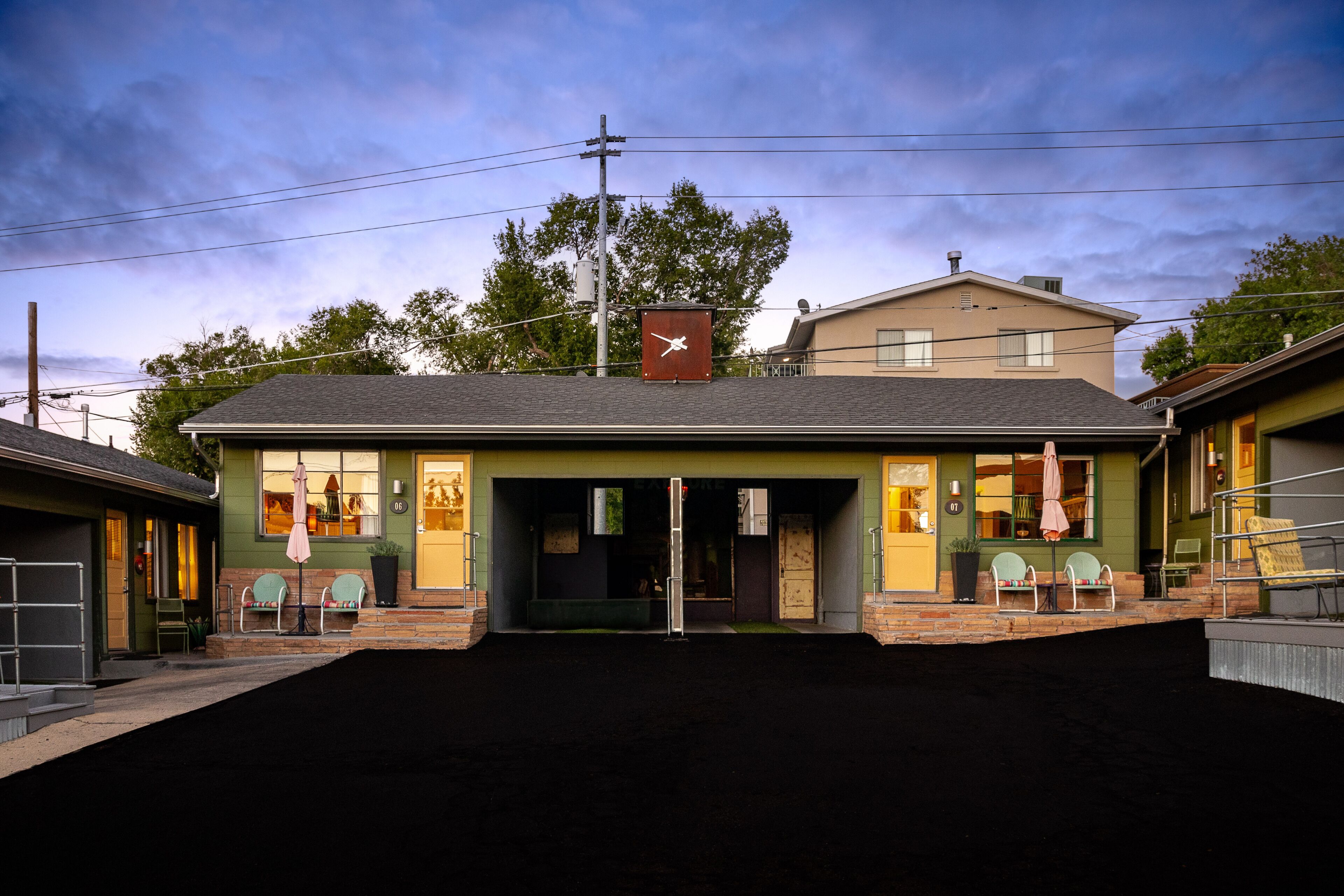 A building with a green facade, a clock on the roof, and a small outdoor seating area with chairs and an umbrella.