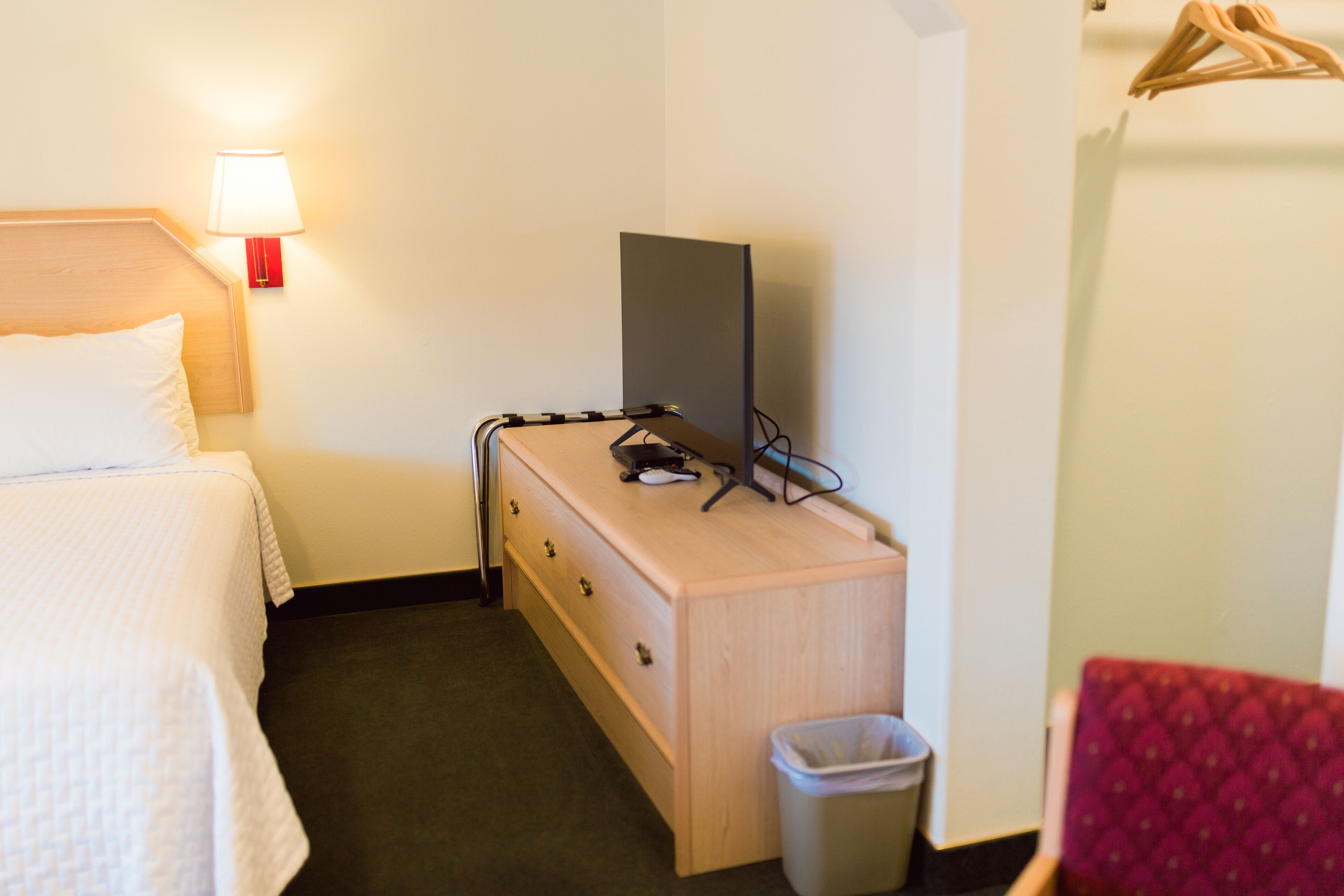 A hotel room with a bed, a wooden dresser, a TV, a lamp, and a trash bin.