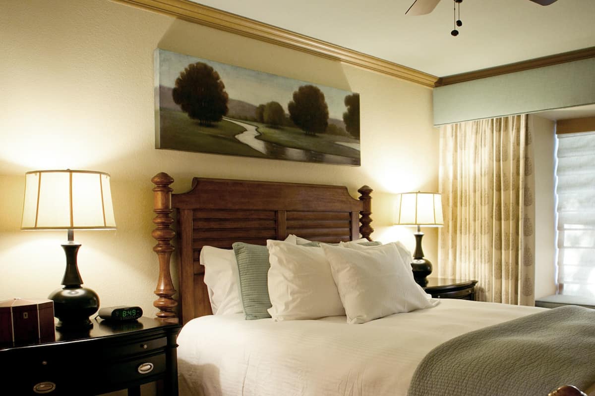 A hotel room with a large bed, two bedside tables, a painting on the wall, and a window with curtains.
