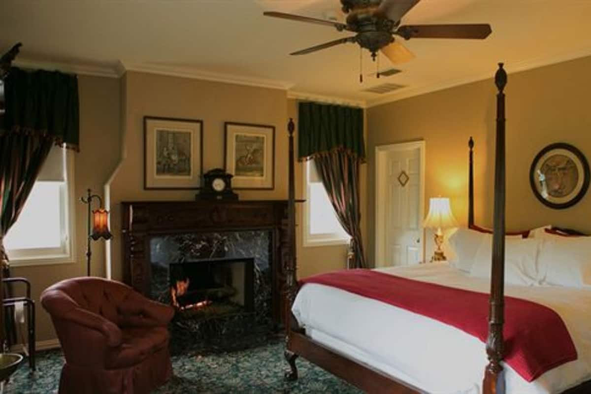 A four-poster bed with a red throw, a fireplace, and a ceiling fan in a room with two framed pictures on the wall.