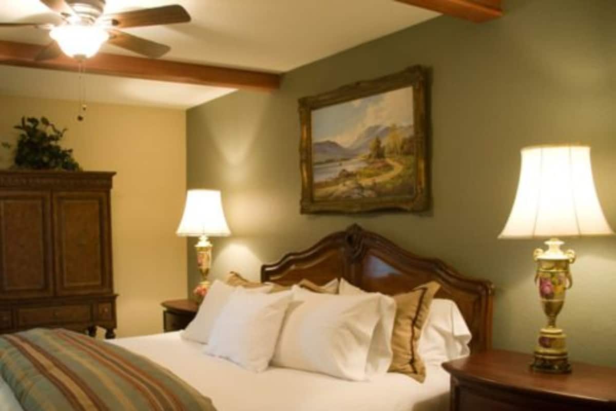 A bedroom with a bed, two bedside lamps, a ceiling fan, a painting on the wall, and a wooden cabinet.