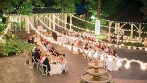 An outdoor event with round tables and chairs, string lights, and a fountain.