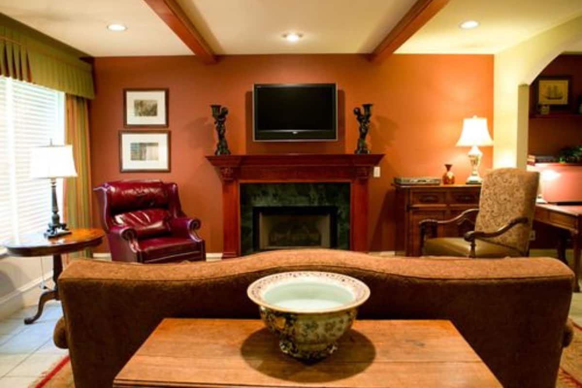 A living room with a fireplace, a television mounted above it, a red leather armchair, a patterned armchair, and a table with a decorative bowl.