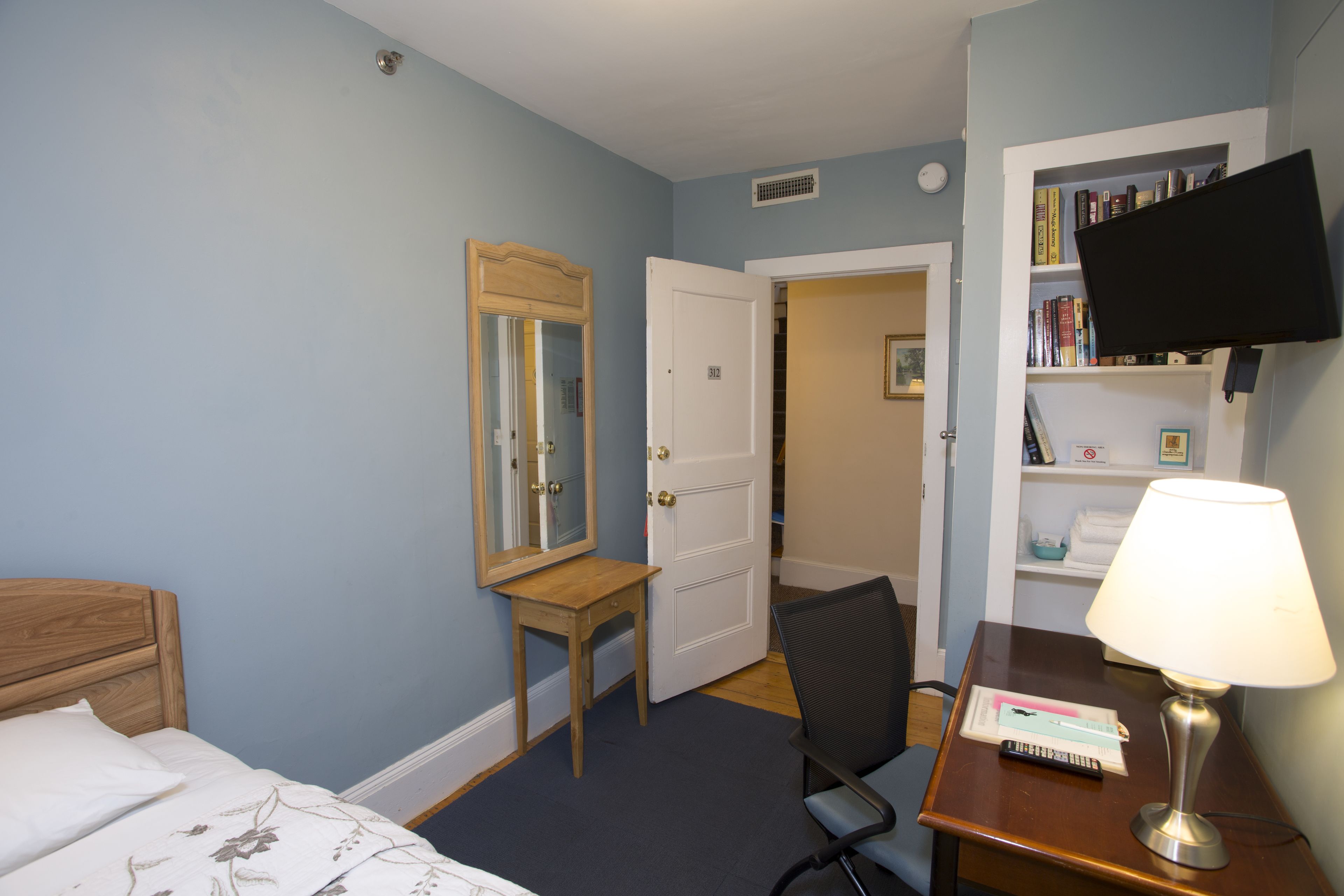 A hotel room with a bed, a desk with a lamp, a chair, a bookshelf, and a television.