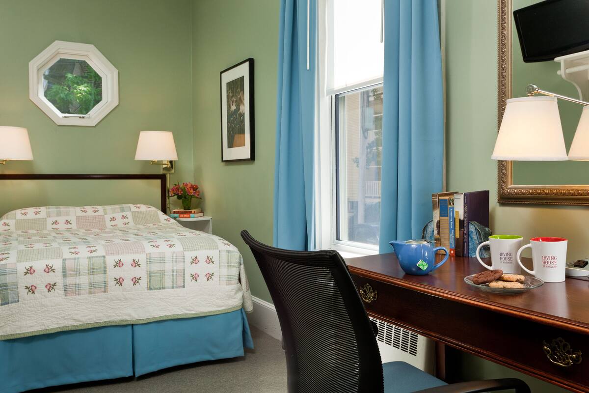 Habitación de hotel con cama, escritorio, silla, ventana con cortinas, televisión y un cuadro en la pared.