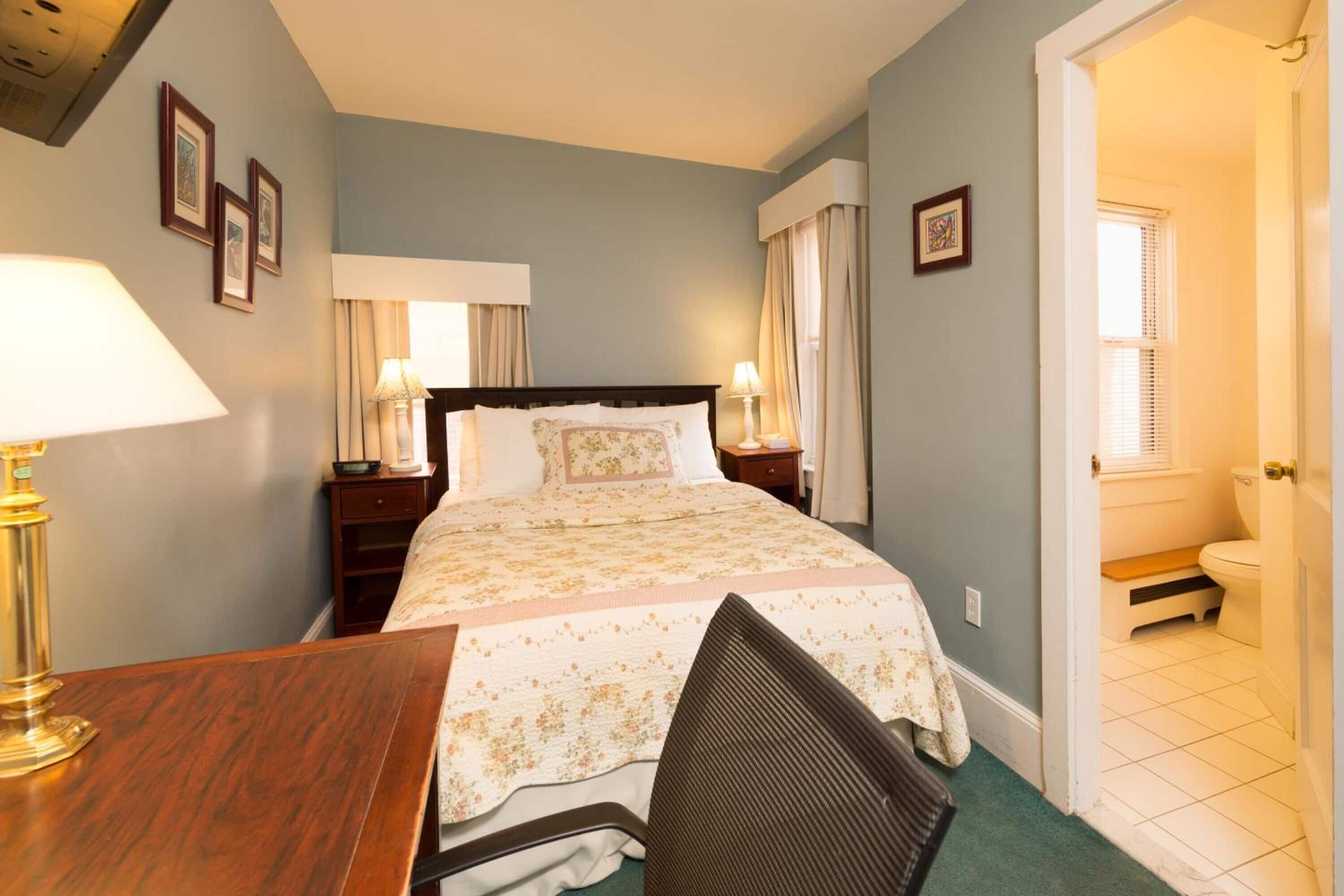 A bedroom with a bed, bedside lamps, a desk, and a chair. There is a bathroom visible through an open door.