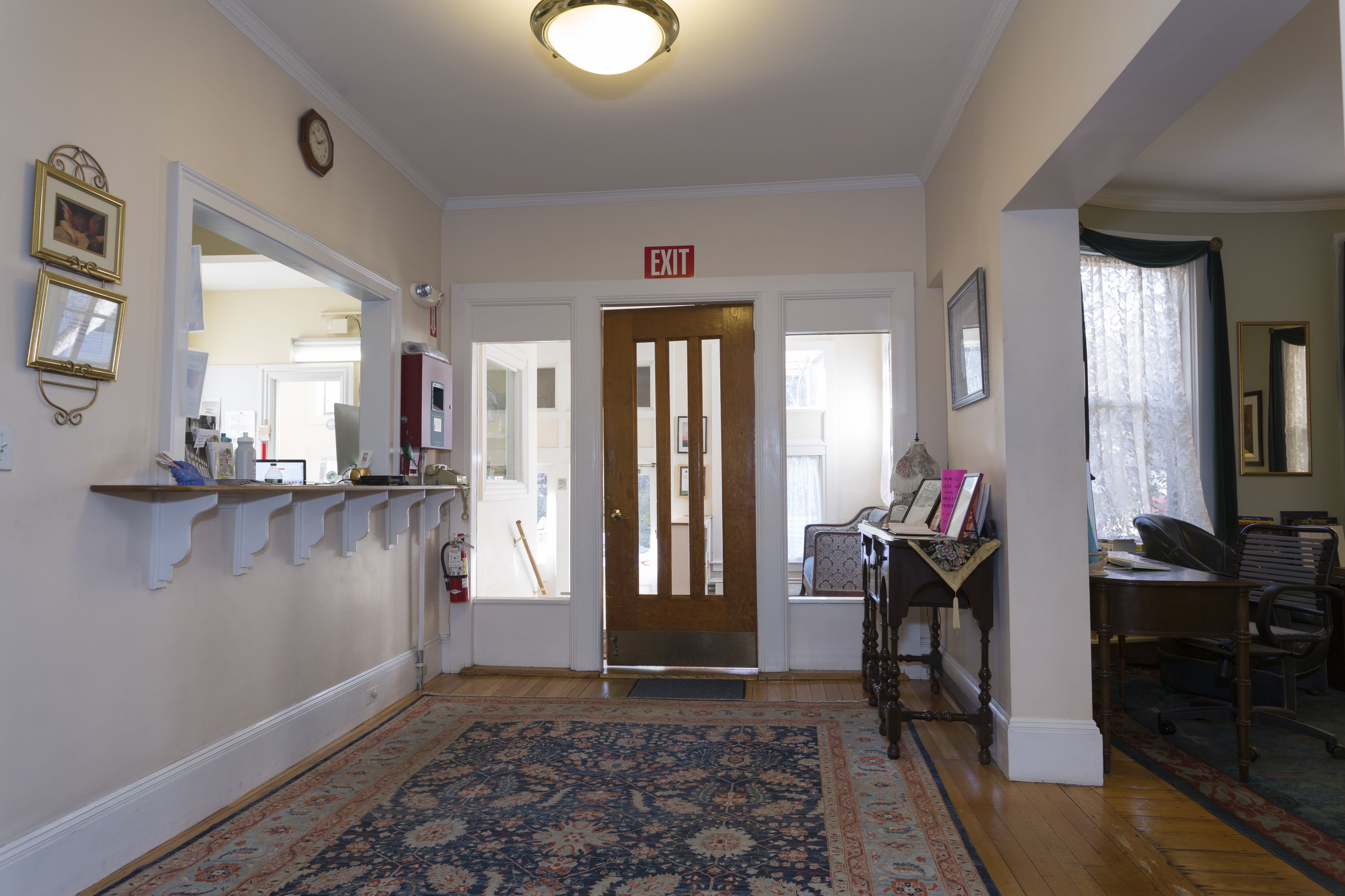 A hallway with a patterned rug, a wooden door with an exit sign, a mirror, and a shelf with various items.