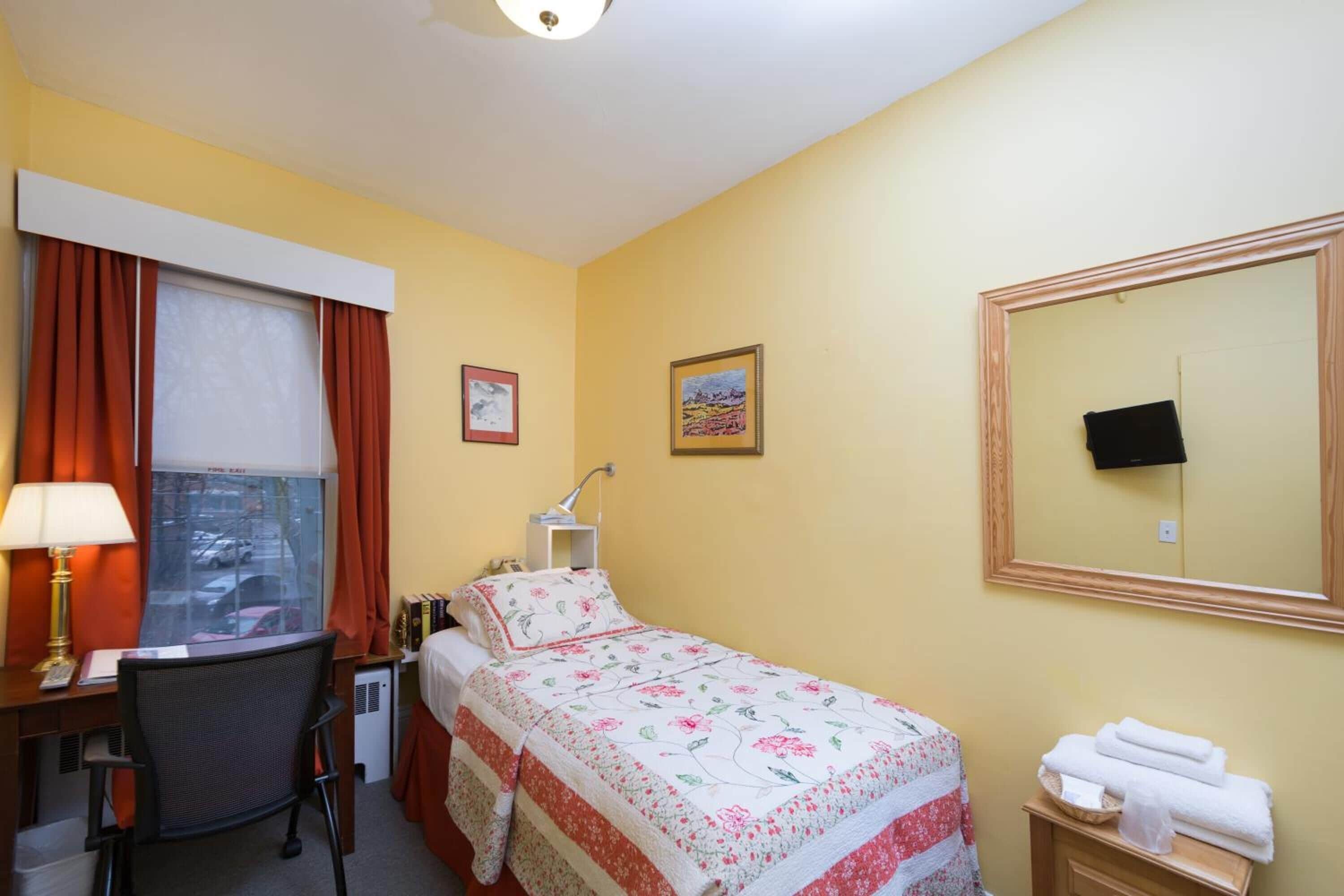 Una habitación de hotel con una cama, un escritorio con lámpara, una silla, un ventanal con cortinas, una mesita de noche con toallas y un espejo en la pared.