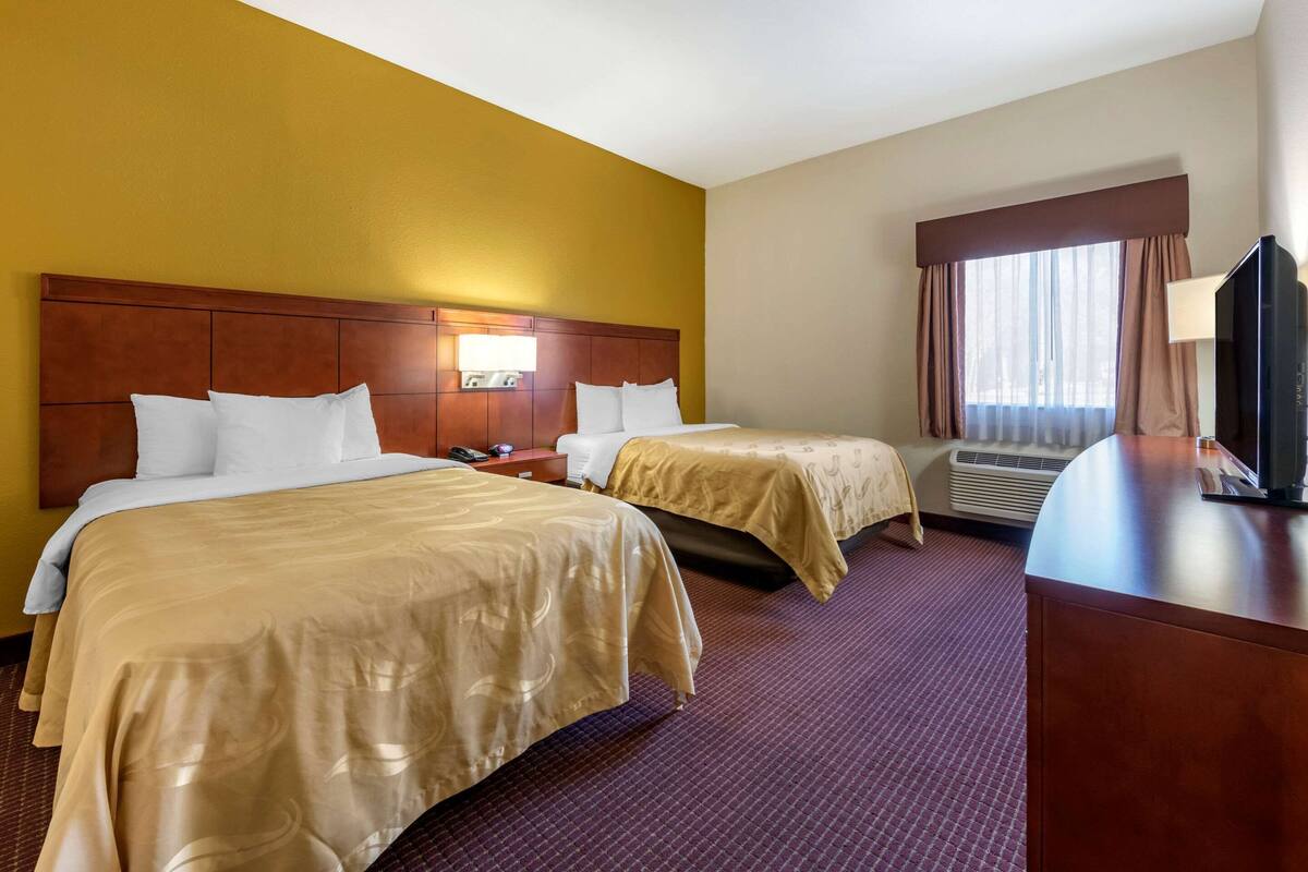 A hotel room with two beds, a wooden headboard, a flat-screen TV, an air conditioning unit, and a window with curtains.