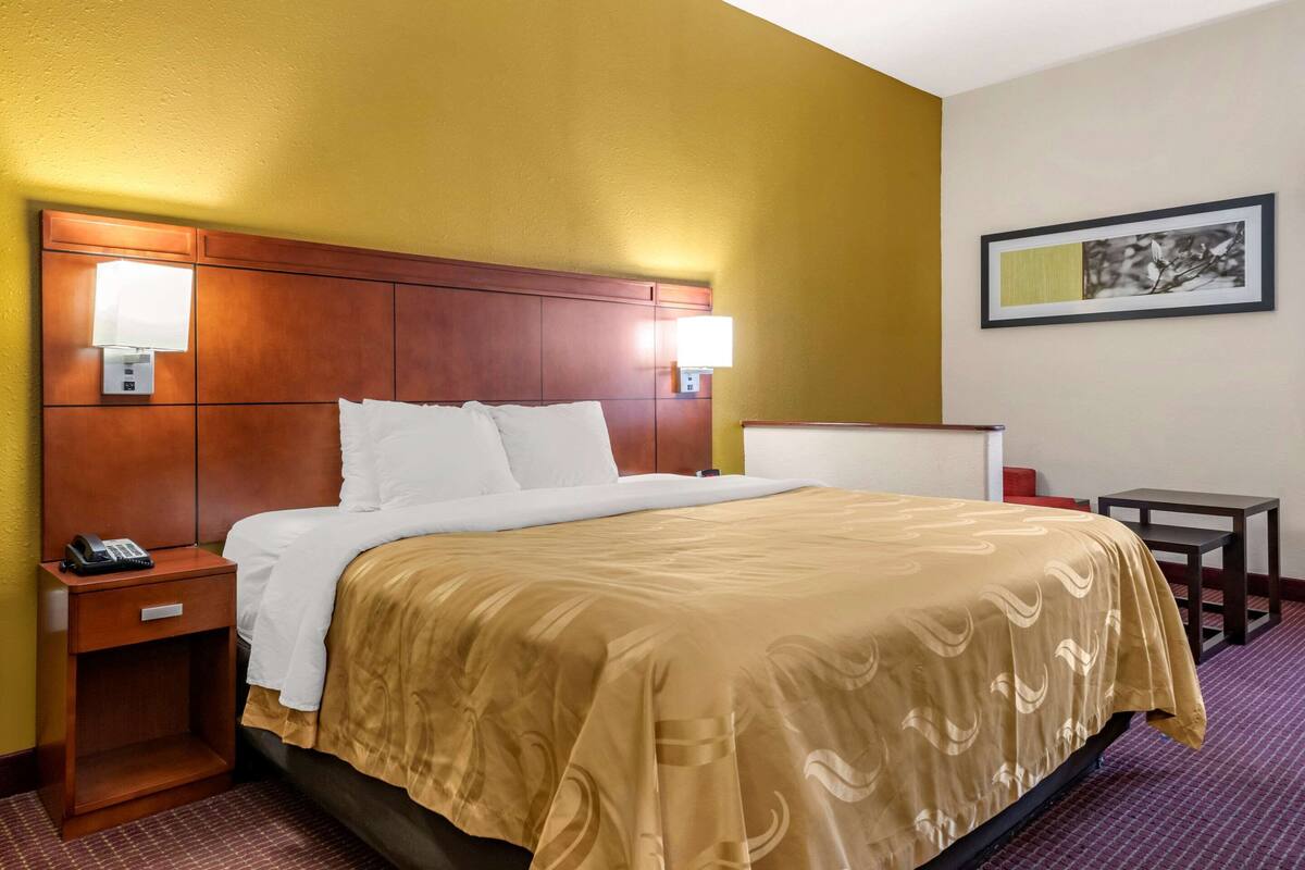 A hotel room with a large bed, wooden headboard, bedside lamps, a small table, and a framed picture on the wall.