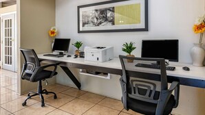 A modern office space with two desks, each equipped with a computer monitor, keyboard, and mouse. There are two office chairs, a printer, and two potted plants on the desks. A framed artwork hangs on the wall above the desks. - Quality Inn Zephyrhills-dade City (Zephyrhills)
