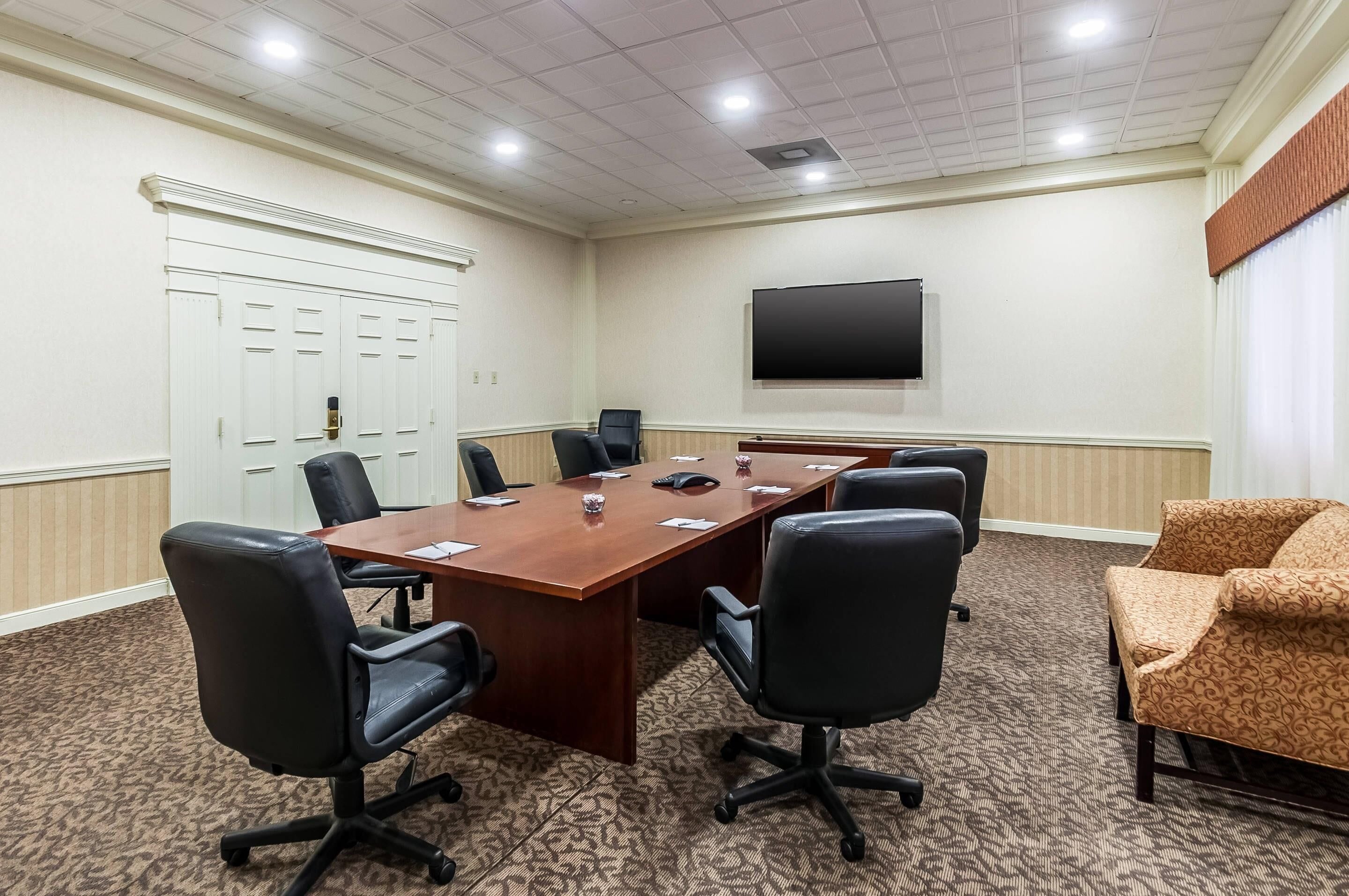 A conference room with a long table, six black office chairs, a television on the wall, and a patterned carpet.