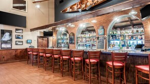 Una zona de bar con sillas de madera, un muro de ladrillo y una variedad de botellas y vasos en estantes.