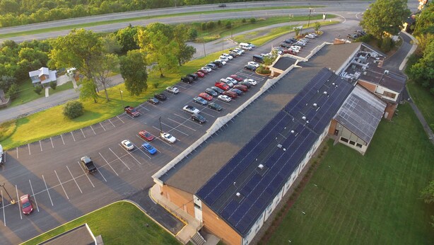 An aerial view of a parking lot with multiple rows of parked cars, a building with a sloped roof, and surrounding greenery. - Clarion Inn Harpers Ferry - Charles Town (Harpers Ferry)
