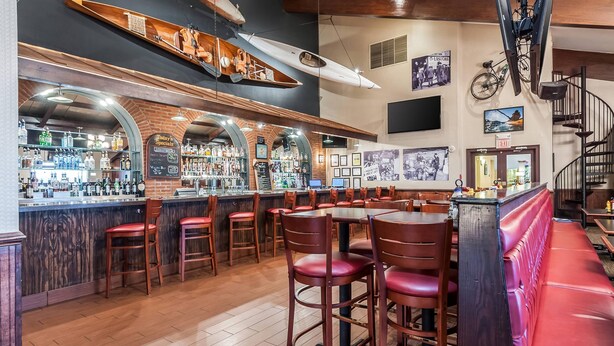 Zona de bar con mostradores de madera, taburetes rojos y un banco rojo. Hay estantes con botellas y una pizarra con la carta.