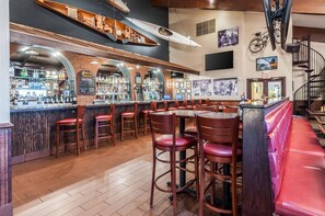 Zona de bar con mostradores de madera, taburetes rojos y un banco rojo. Hay estantes con botellas y una pizarra con la carta.