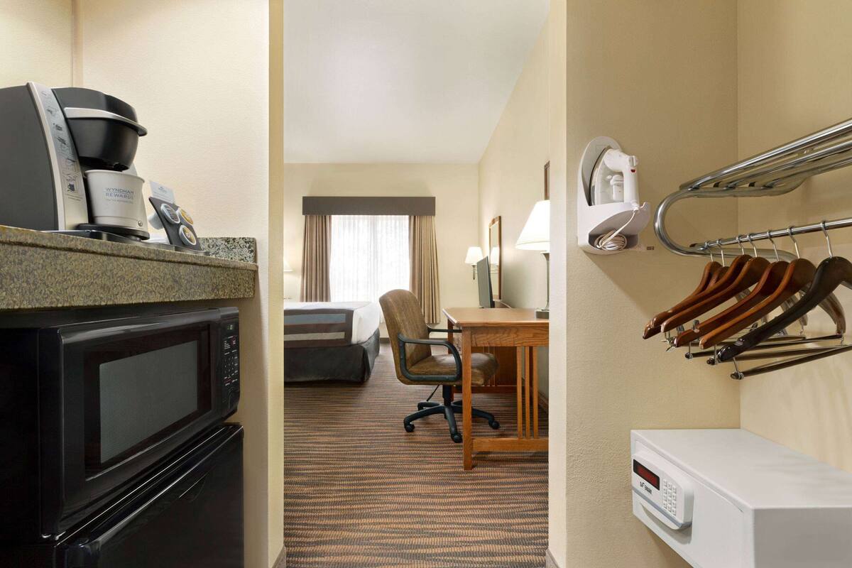 A hotel room with a kitchenette including a microwave, coffee maker, and iron. There is a desk with a lamp, a chair, and a rack with irons. The room has a carpeted floor and a view through a window with curtains.