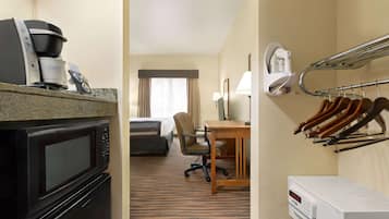 A hotel room with a kitchenette including a microwave, coffee maker, and iron. There is a desk with a lamp, a chair, and a rack with irons. The room has a carpeted floor and a view through a window with curtains.