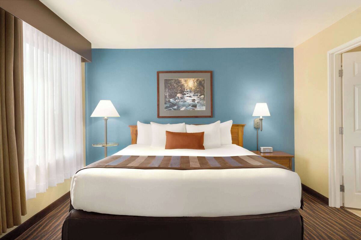 A hotel room with a large bed, two bedside lamps, a framed picture on the wall, and a window with curtains.