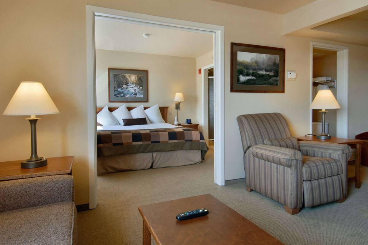 A hotel room with a bed, a chair, a table, and a remote control.