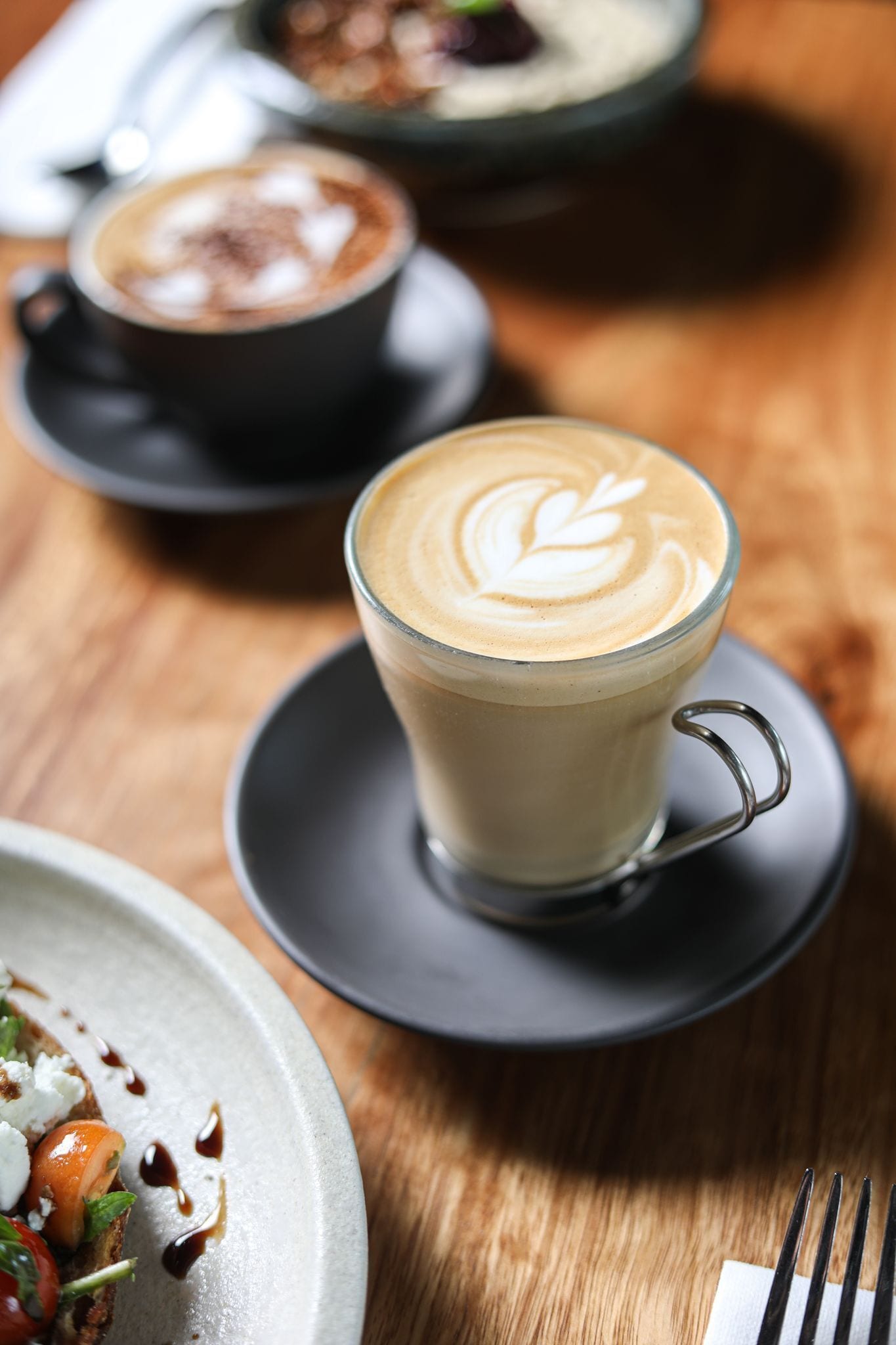 A table with a glass cup of coffee with latte art, a plate with food, and another cup of coffee in the background.