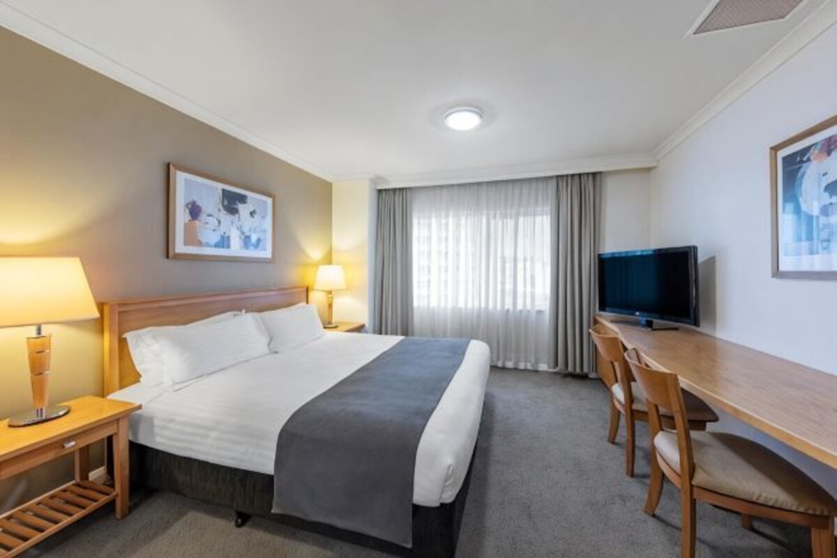 A hotel room with a bed, bedside tables, a desk with a chair, a television, and a window with curtains.