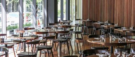 A spacious restaurant with high ceilings and large windows, offering a view of the outdoors. Tables are neatly arranged with chairs, and the interior features a modern design with wooden accents.
