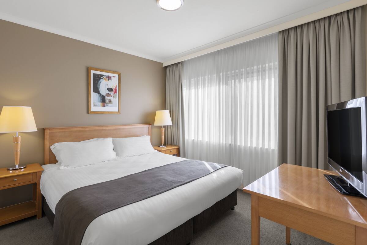 Habitación de hotel con una cama grande, dos lámparas de noche, un televisor, un escritorio y una ventana con cortinas.