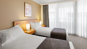 Hotelkamer met twee bedden, een nachtkastje met lamp, een lijst met foto aan de muur en een groot raam met dunne gordijnen.