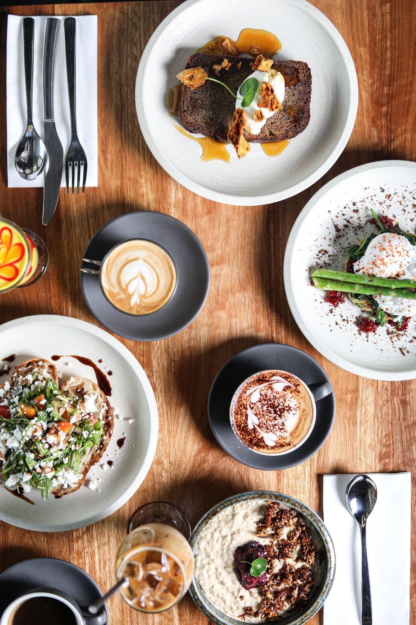 A variety of dishes on a wooden table, including a plate with a dark bread topped with sauce and nuts, another with a green vegetable and sauce, and a bowl of creamy food with a berry.