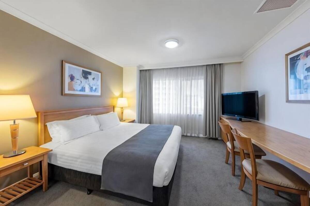 Habitación de hotel con cama, mesitas de noche, escritorio con silla, televisor y ventana con cortinas.