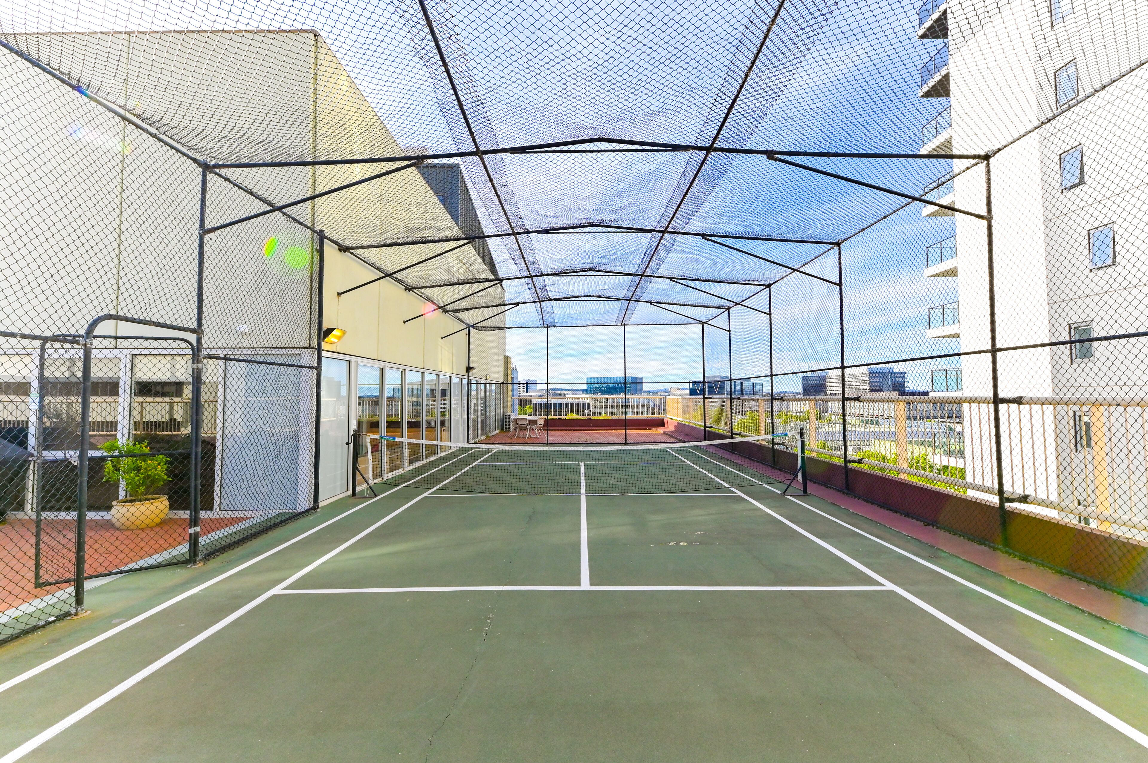 An indoor tennis court with a net, surrounded by a metal fence and glass walls, with a view of buildings in the background.