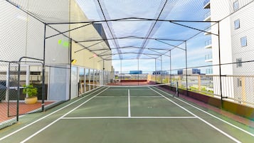 Una cancha de tenis cubierta con red, rodeada por una valla metálica y paredes de vidrio, con vistas a edificios al fondo.