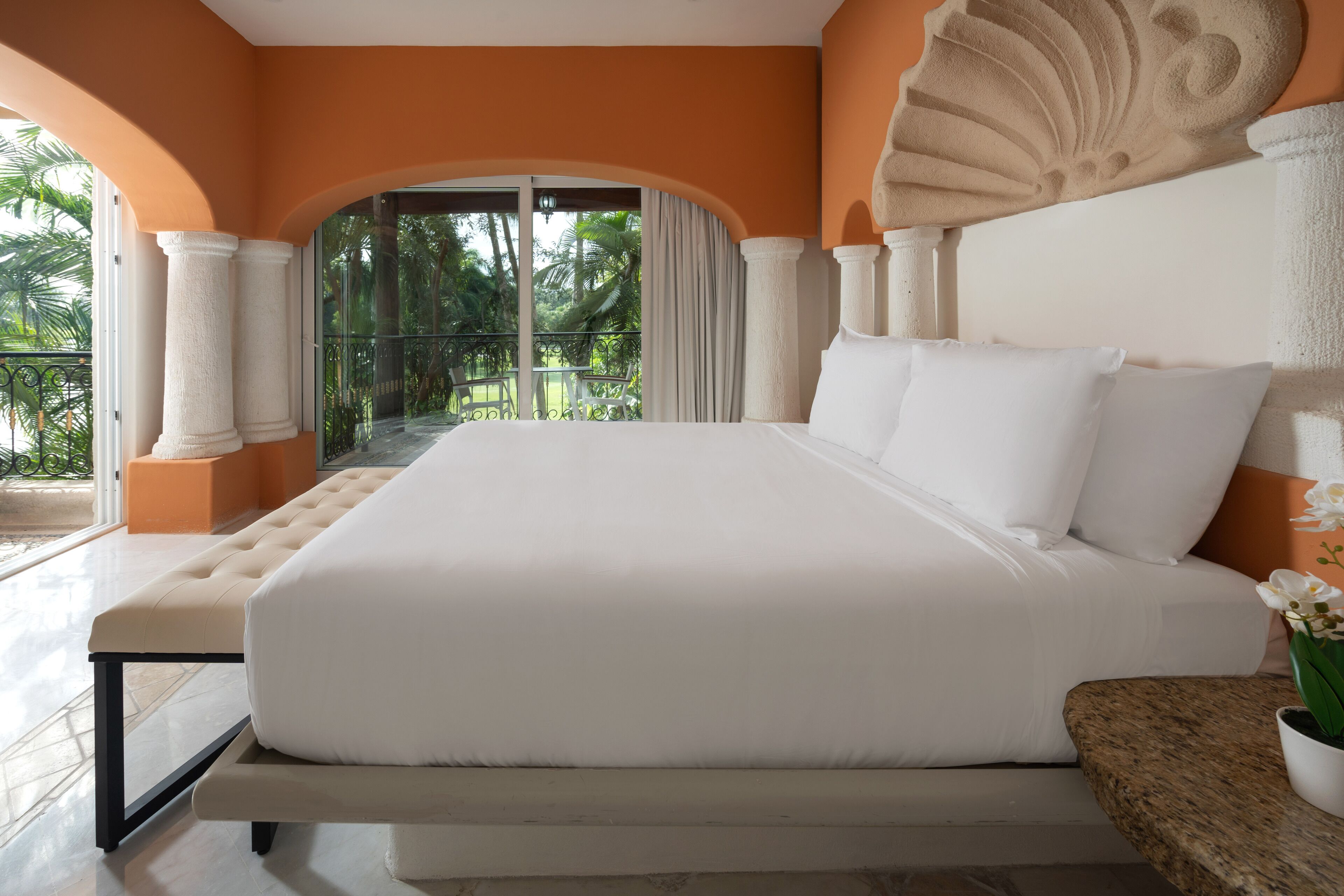 Una cama grande con sábanas blancas, un banco y una planta en maceta en una habitación con paredes anaranjadas y un balcón con vistas a palmeras.