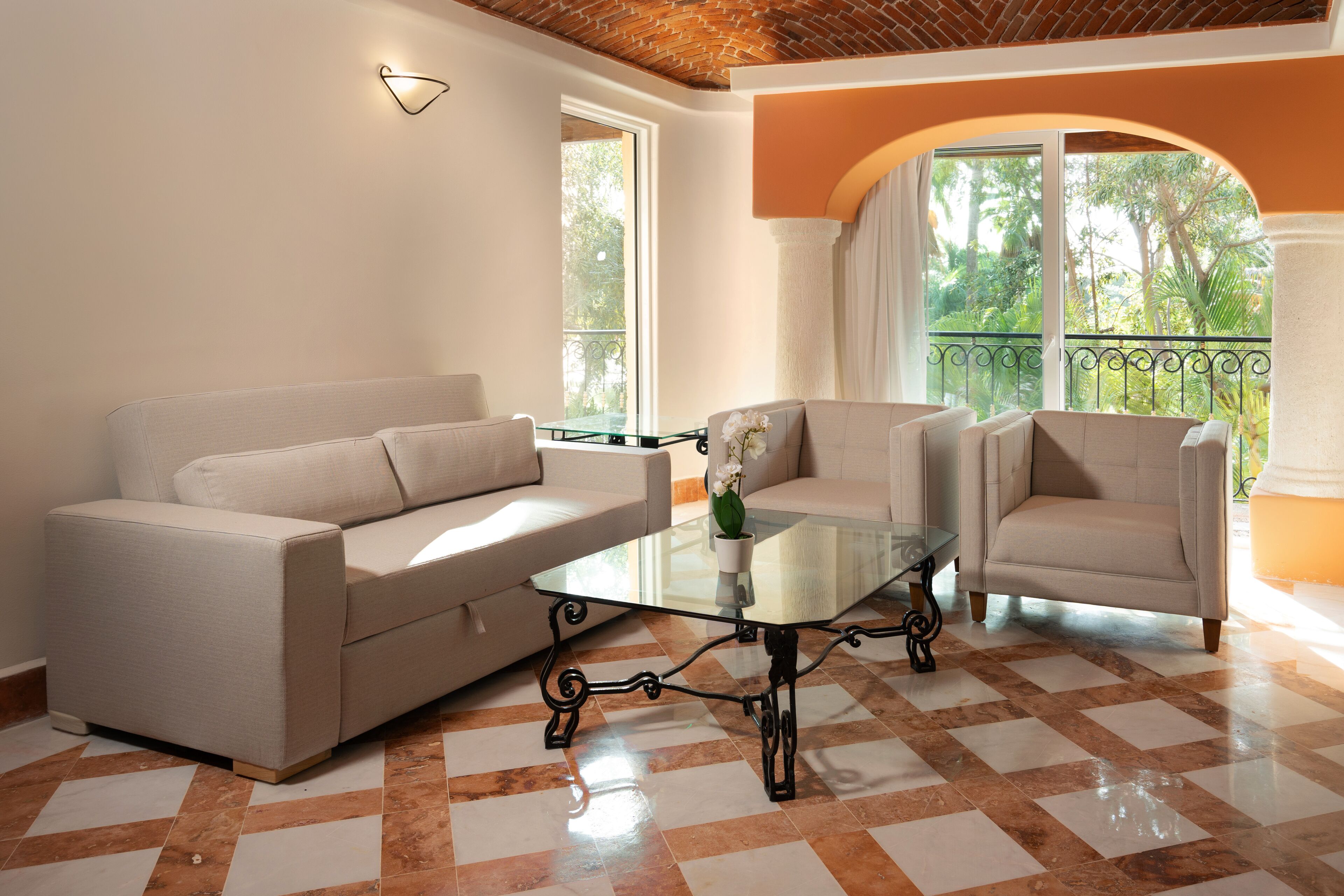 A living room with a beige sofa, two armchairs, a glass coffee table, and a view of greenery through large windows.
