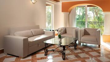 A living room with a beige sofa, two armchairs, a glass coffee table, and a view of greenery through large windows.