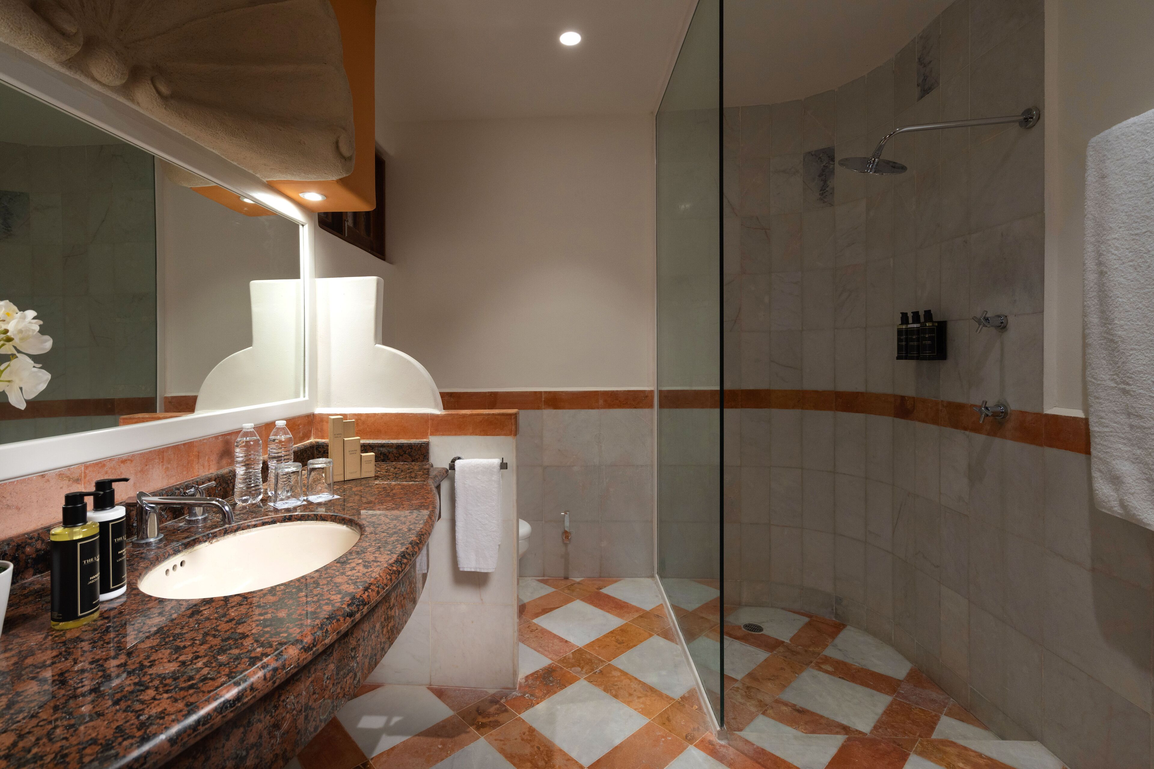 Ein Badezimmer mit einer Granitplatte, einem Waschbecken, einem Spiegel, einer Dusche mit Glastür und einem gefliesten Boden.