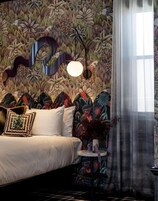 Una camera da letto con carta da parati a motivi floreali, un letto con lenzuola bianche, un tavolino rotondo con un vaso e una finestra con tende leggere.