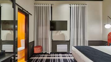 A hotel room with a bed, a flat-screen TV mounted on the wall, a heater, a small round ottoman, and a window with curtains.