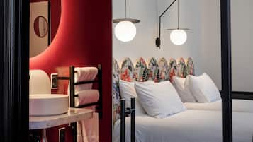 Un baño moderno con una pared roja de acento, un espejo redondo y un lavabo blanco. La habitación tiene un piso a cuadros blanco y negro y una cama con ropa de cama blanca y un cabecero floral.