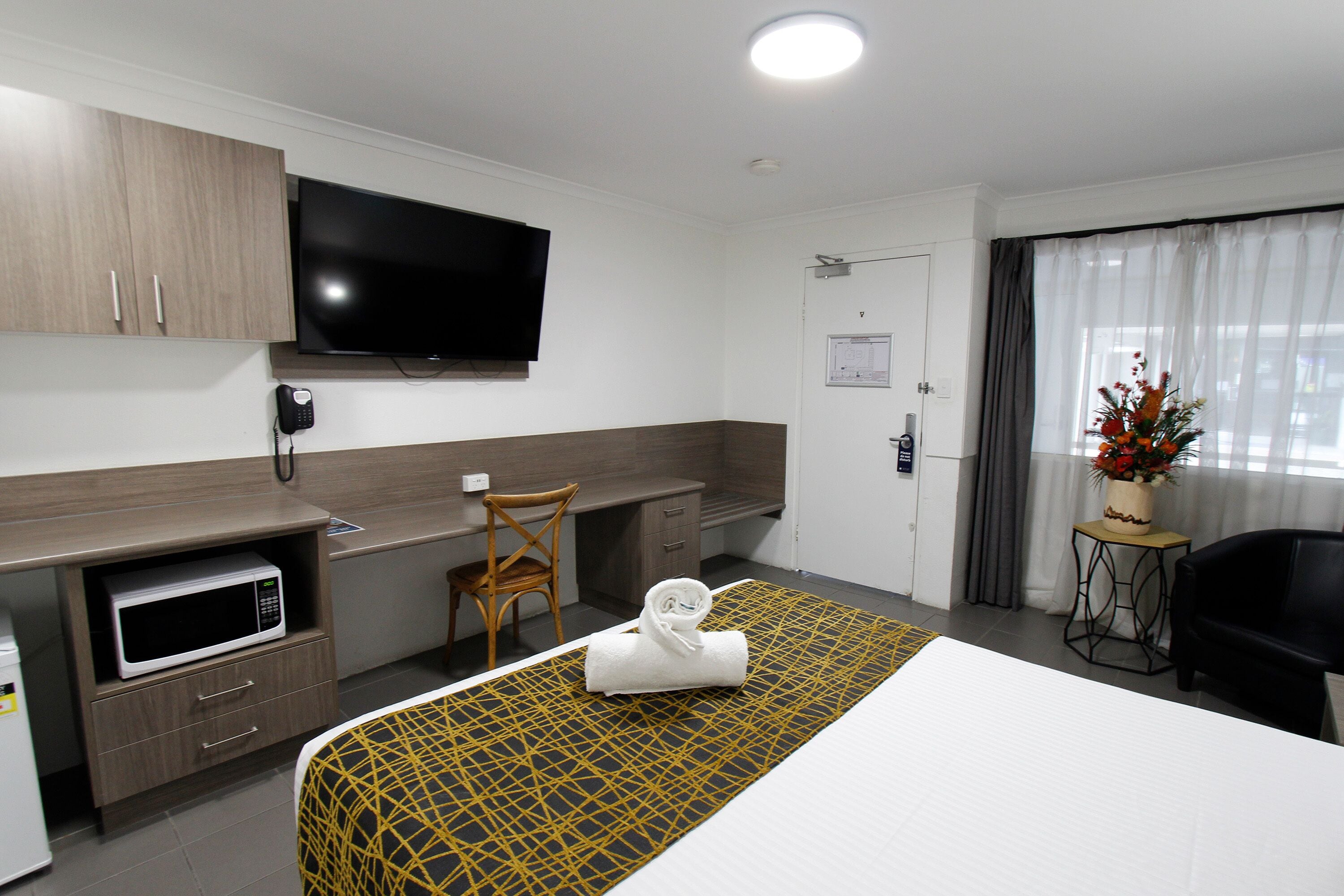 Habitación de hotel con cama, microondas, televisión, escritorio, silla y una pequeña mesa con un jarrón de flores.