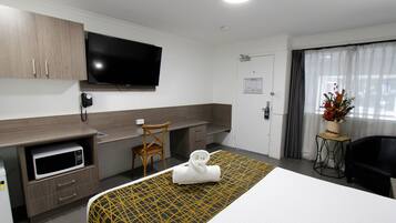 Habitación de hotel con cama, microondas, televisión, escritorio, silla y una pequeña mesa con un jarrón de flores.
