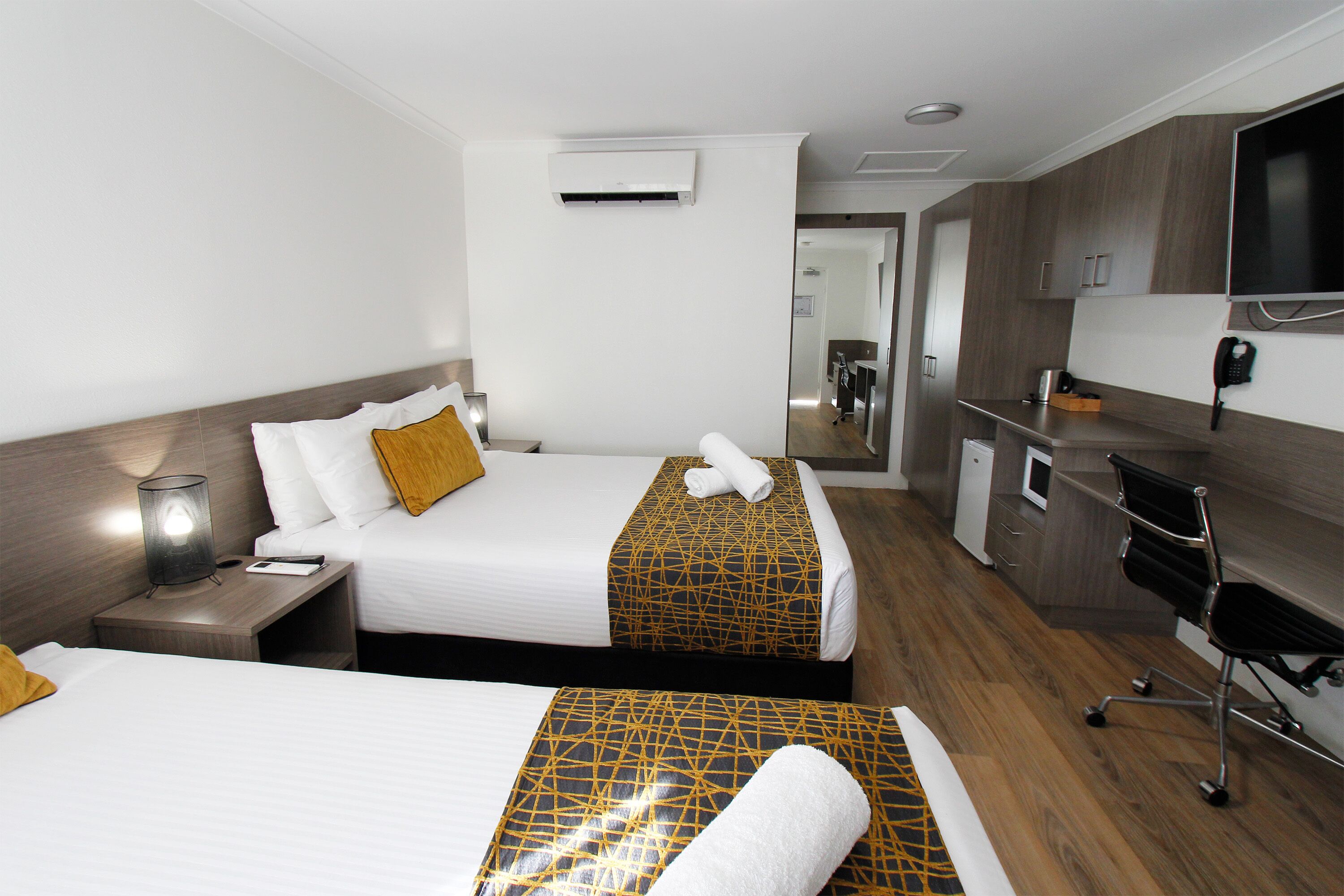A hotel room with two beds, a desk, a chair, a TV, and a kitchenette.