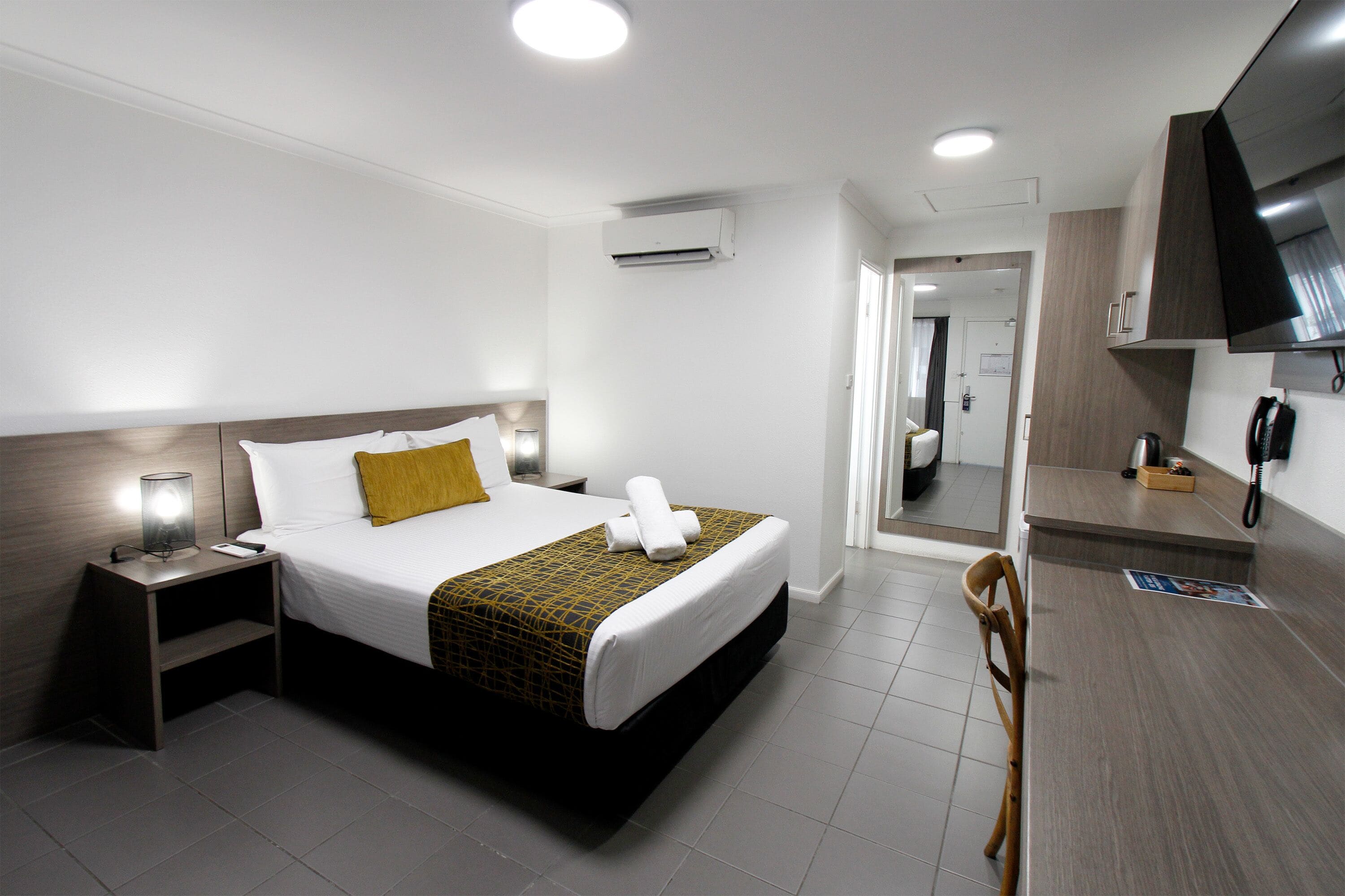 Quarto de hotel com cama, mesa de cabeceira, abajur e TV fixada na parede.