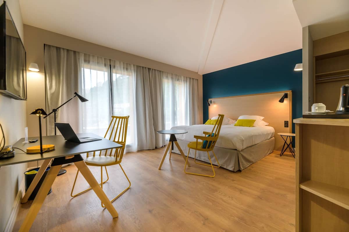 A hotel room with a bed, a desk with a laptop, two chairs, a small table, and a nightstand.