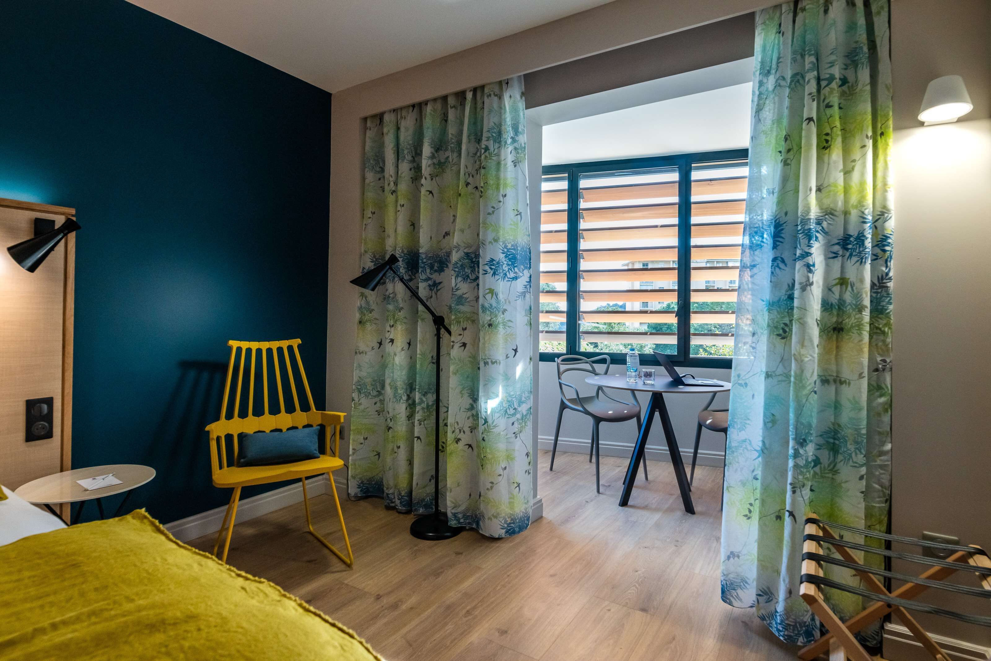 A modern room with a bed, a yellow chair, a table with chairs, a floor lamp, and a window with blinds.