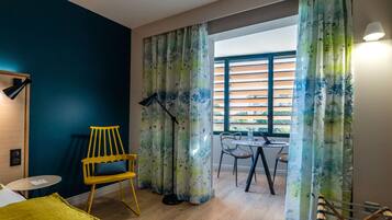 A modern room with a bed, a yellow chair, a table with chairs, a floor lamp, and a window with blinds.